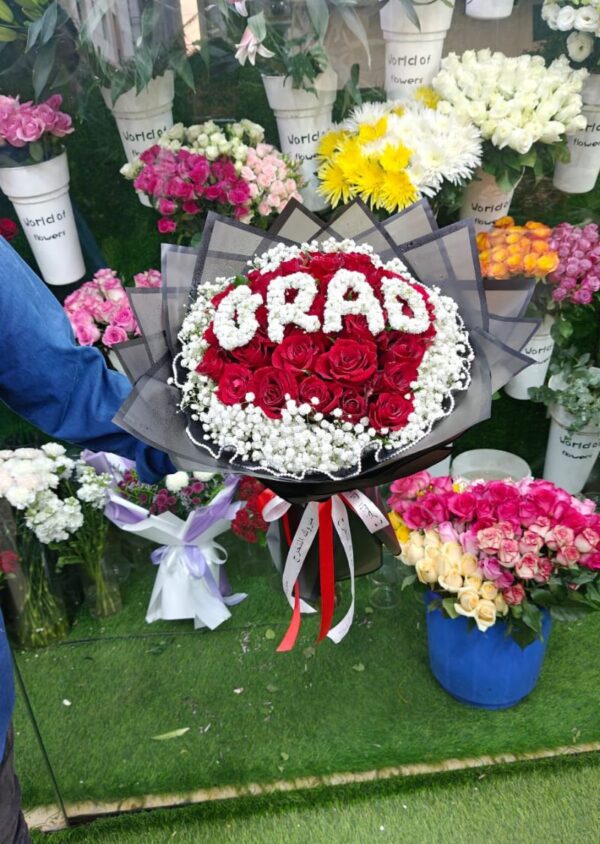 Graduation bouquet of red roses with 'GRAD' lettering in white flowers, wrapped in black paper, perfect for graduation gifts in Dubai.