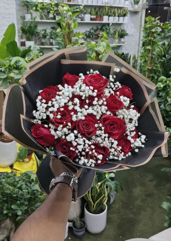 Hand holding a bouquet of fresh red roses with baby’s breath, arranged in black wrapping paper inside a Dubai flower shop.