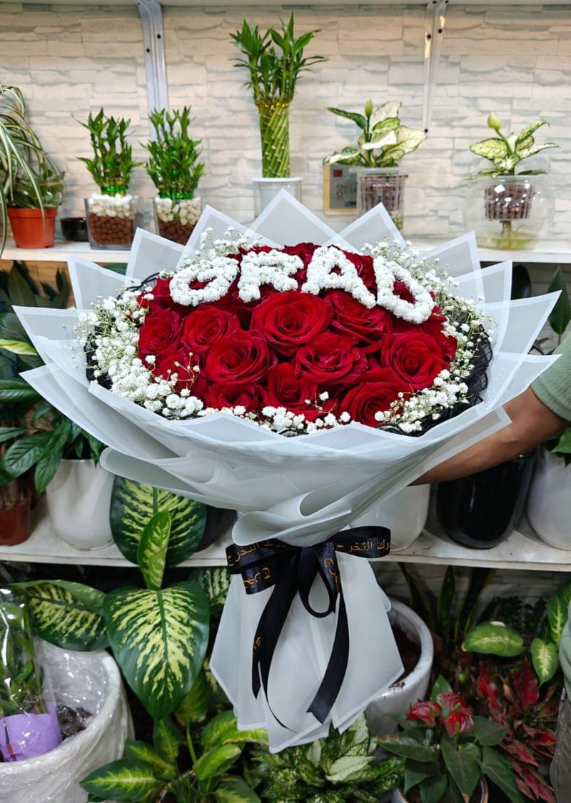 A beautifully handcrafted graduation flower bouquet featuring deep red roses arranged in a round shape, accented with delicate white flowers that spell out "GRAD" — perfect for celebrating academic achievements in the UAE.